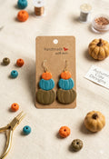 Handmade stacked pumpkin clay earrings in olive, teal, and orange on a kraft paper display card